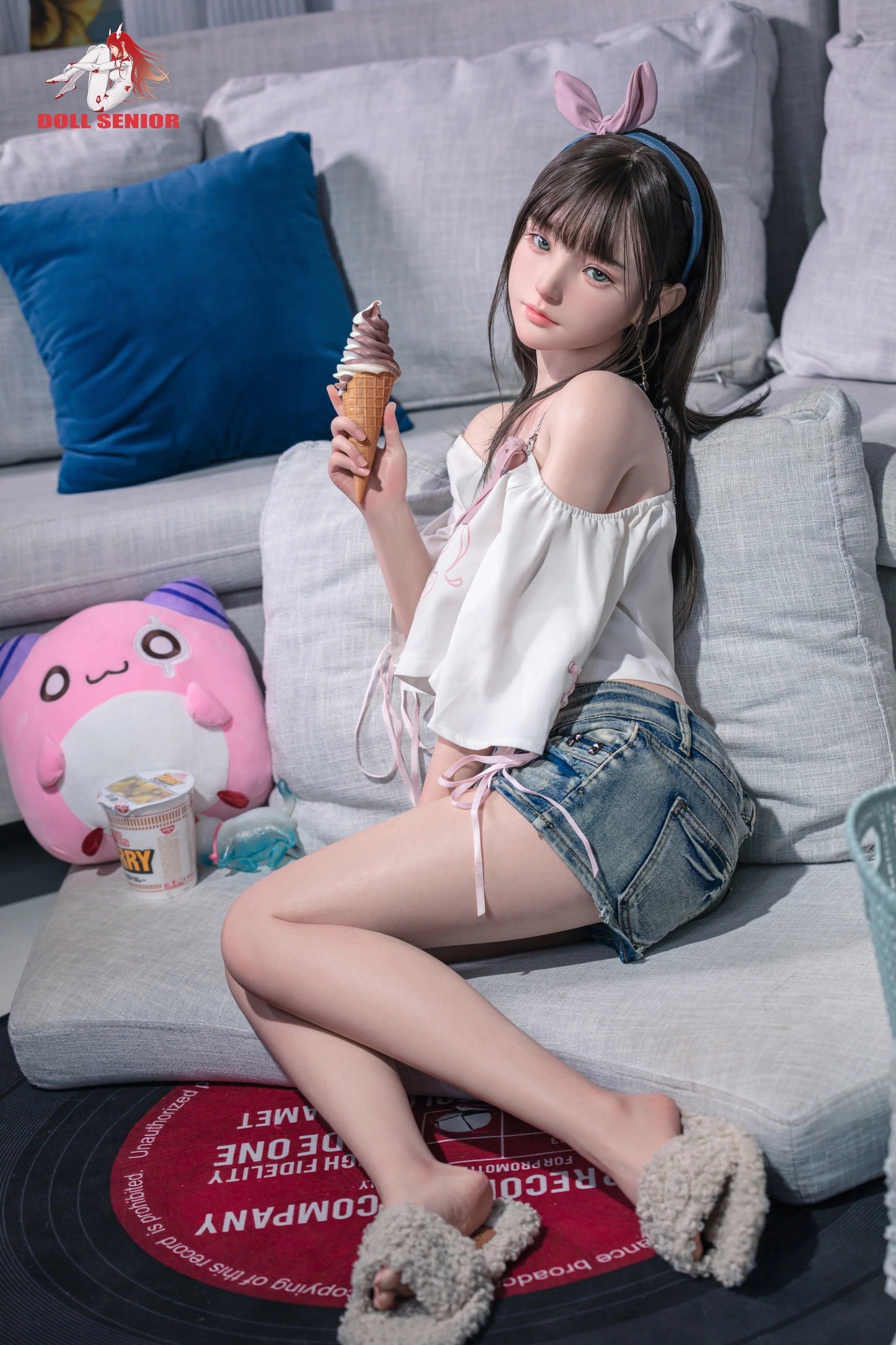 Realistic female doll with brown hair, pink bunny ears, holding ice cream, wearing white top and denim shorts on gray couch