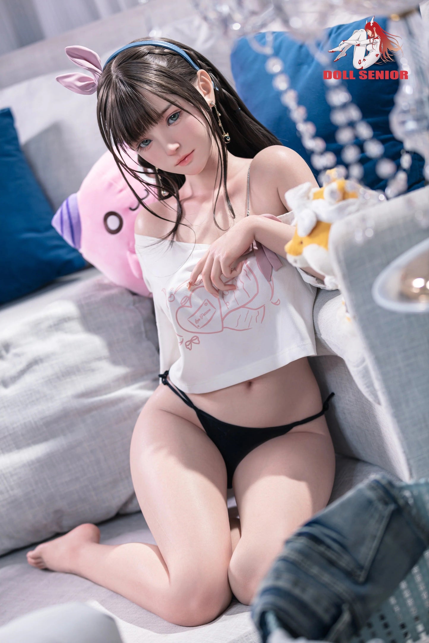 Realistic female doll with brown hair, blue eyes, wearing white top and black underwear, sitting on couch with plush toys.