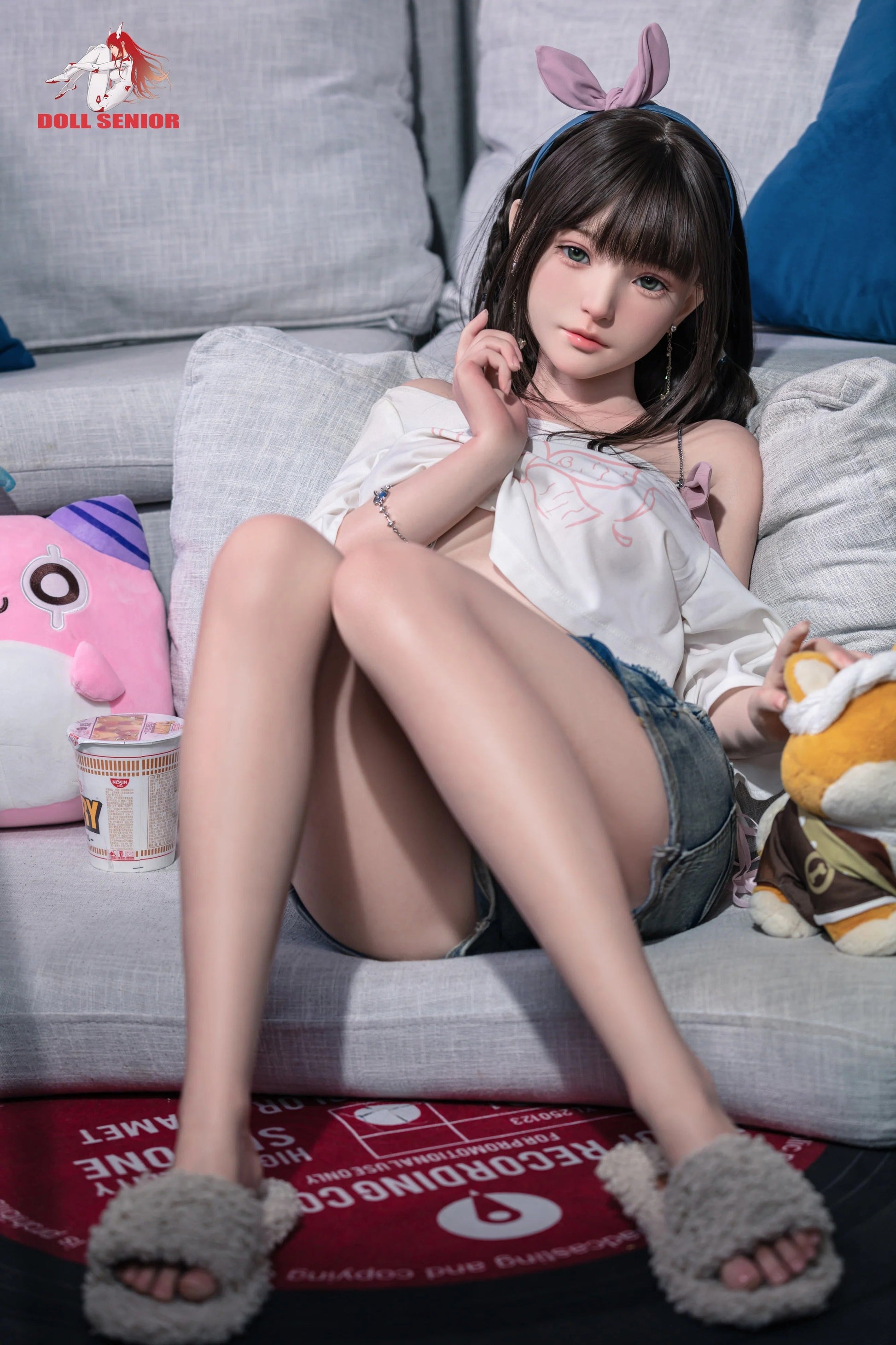 Realistic female doll with brown hair, green eyes, wearing white top, denim shorts, bunny ears, and fuzzy slippers on couch.