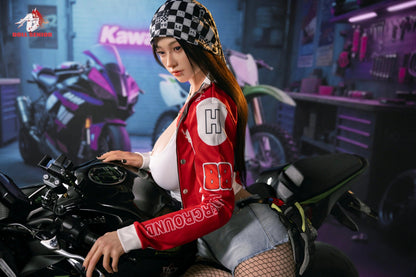 Realistic female doll in red racing jacket and checkered bandana, seated on a motorcycle in a garage setting.