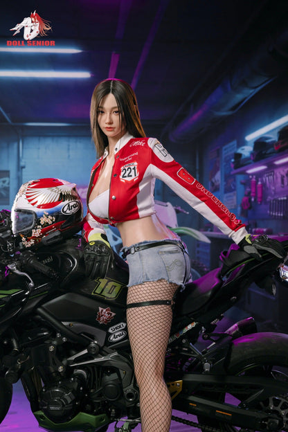 Realistic female doll in red racing jacket and denim shorts posing beside black motorcycle in neon-lit garage