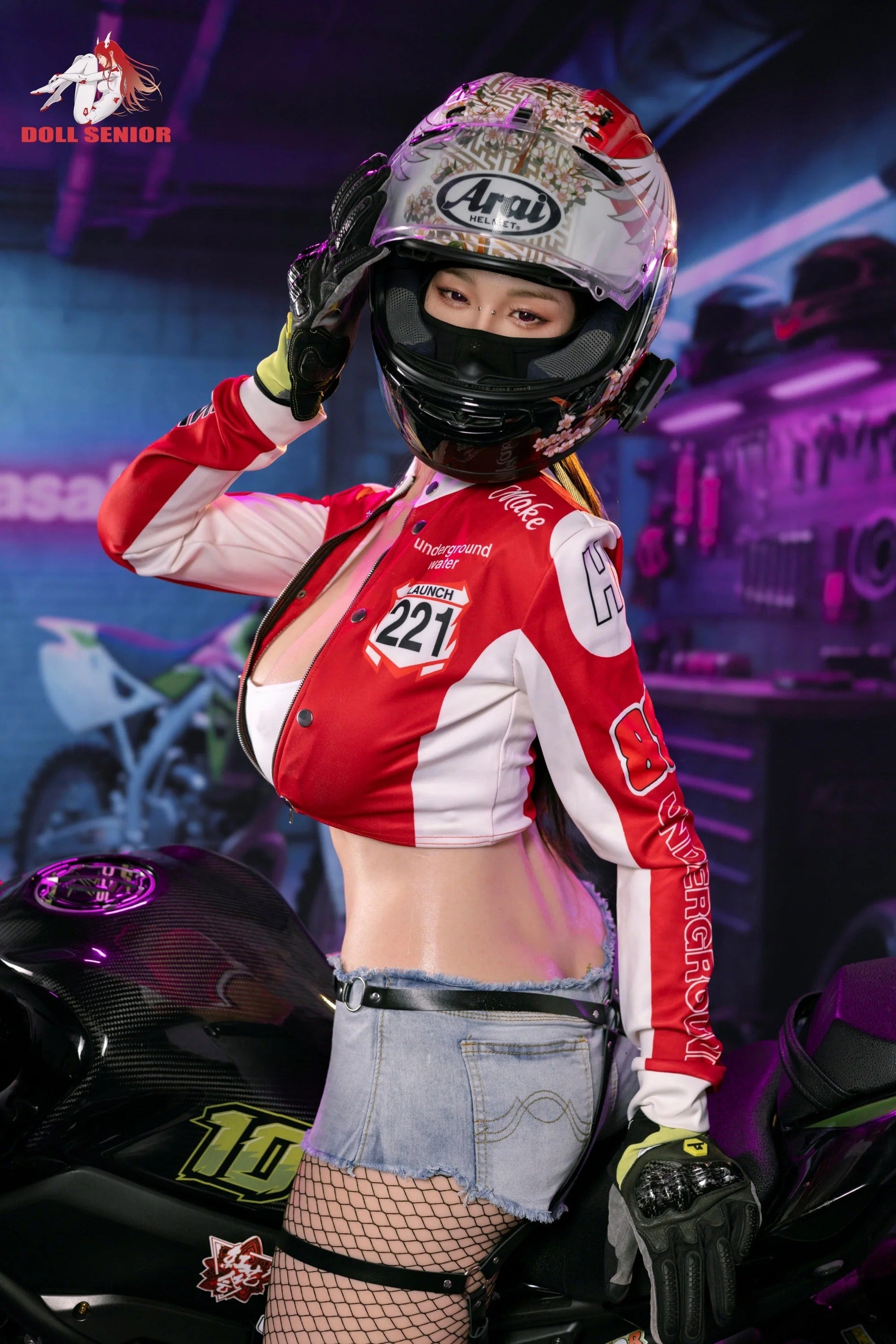 Realistic female doll in red racing jacket, floral helmet, denim shorts, and fishnet stockings beside motorcycle