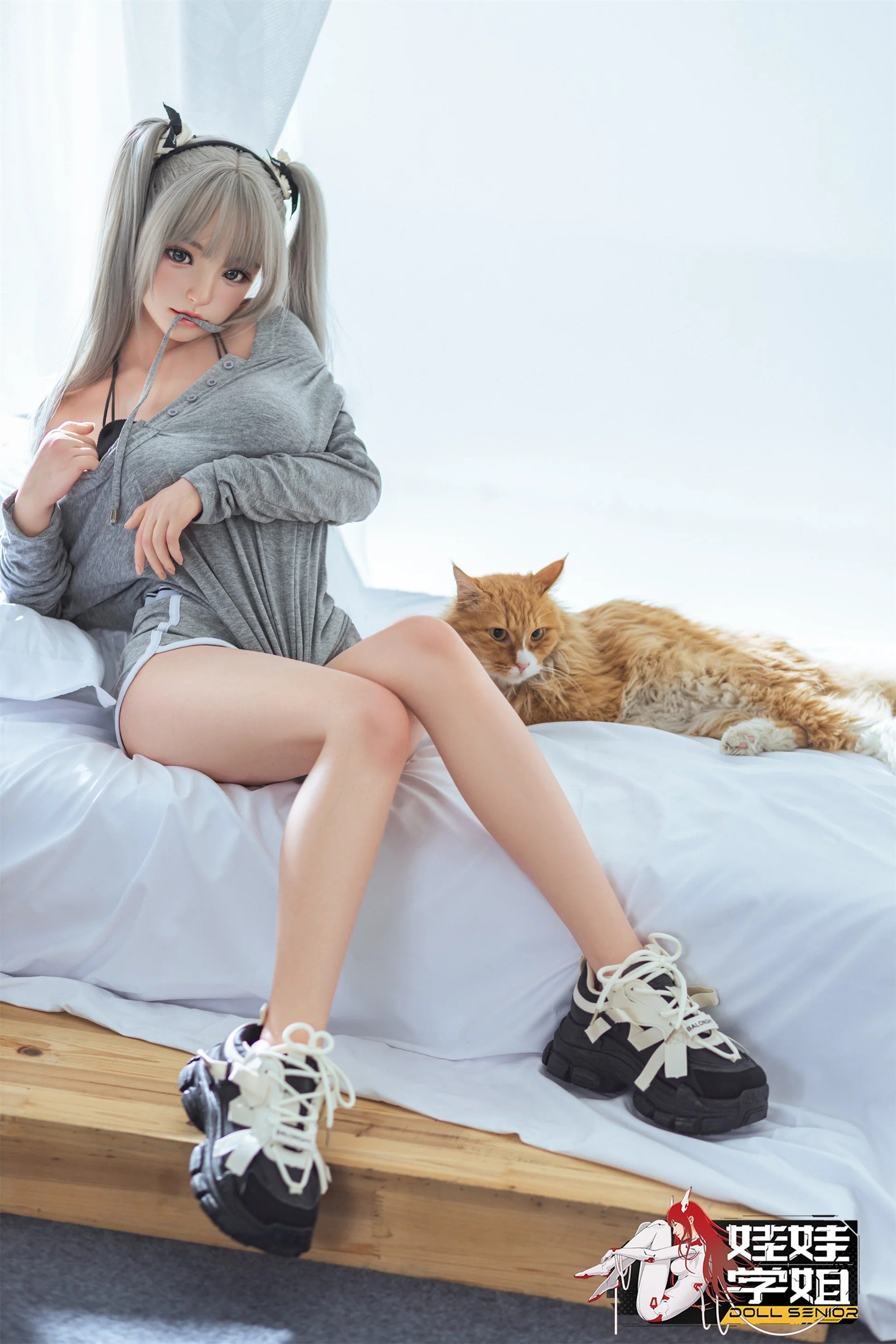 Realistic anime-style doll with silver pigtails, wearing gray hoodie and black sneakers, seated beside ginger cat on white bed.
